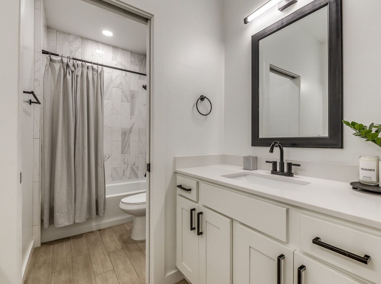 A bathroom with a white shaker vanity and black framed mirror, the tub and shower visible through the doorway