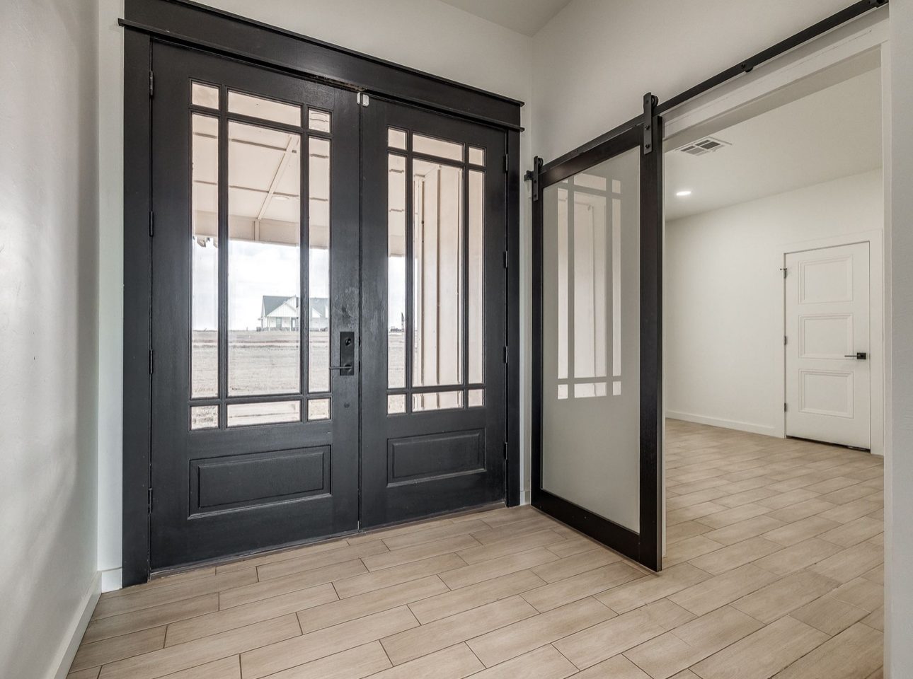 Black double entry doors with divided glass beside a frosted glass barn door on a black track, over plank tile