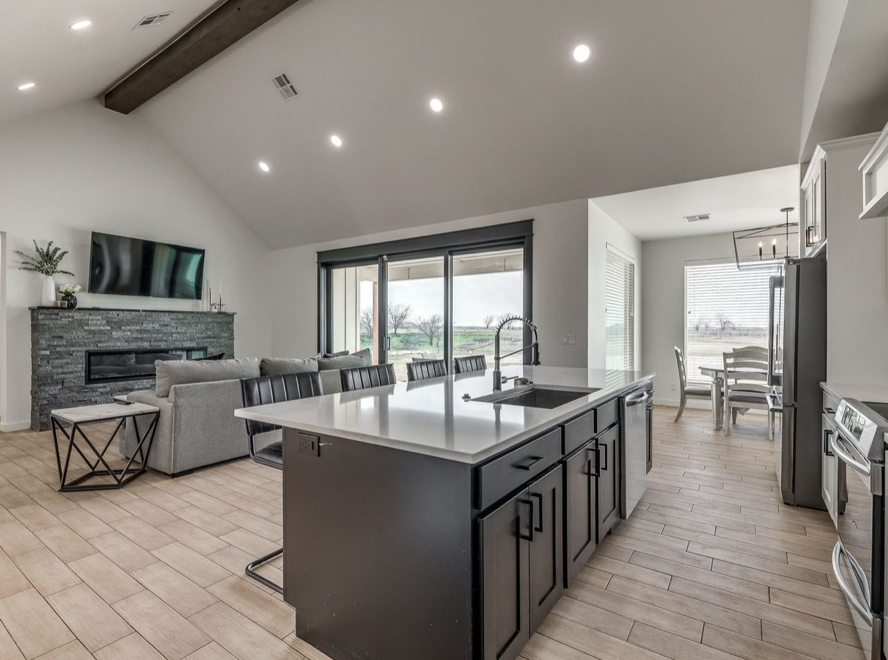 A kitchen with a dark island and white quartz top, stainless appliances and pendant lights, and a stone fireplace in the living room beyond