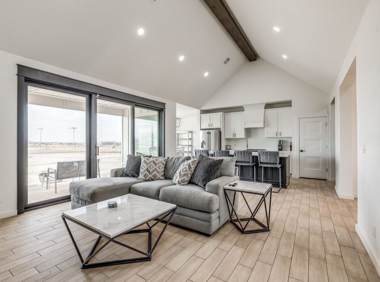 A living room under a vaulted ceiling with an exposed beam, a gray sectional and marble topped table, and sliding doors to the patio