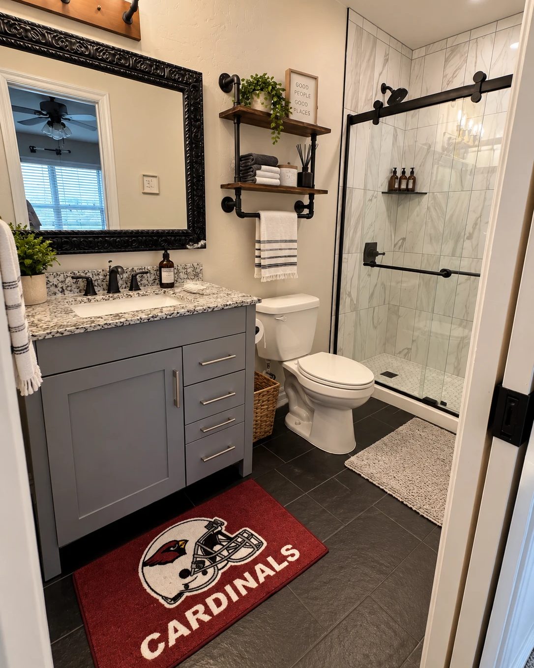 A finished bathroom with a gray shaker vanity under a speckled granite top, a black framed mirror, black pipe shelving holding towels and a plant, a dark slate tile floor, and a marble look tile shower behind a sliding glass door on a black track