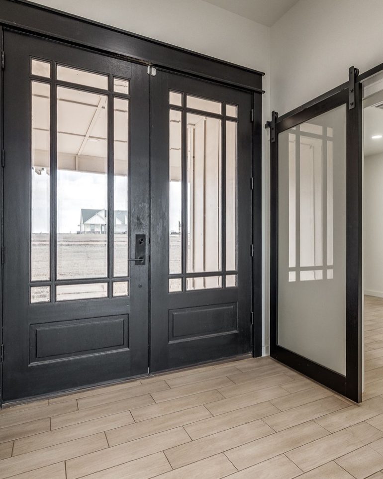 Black double entry doors with divided glass beside a frosted glass barn door on a black track, over plank tile