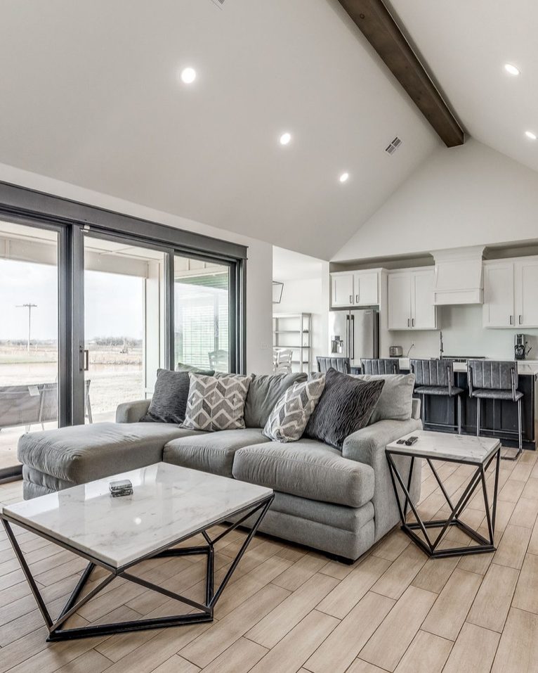 A living room under a vaulted ceiling with an exposed beam, a gray sectional and marble topped table, and sliding doors to the patio