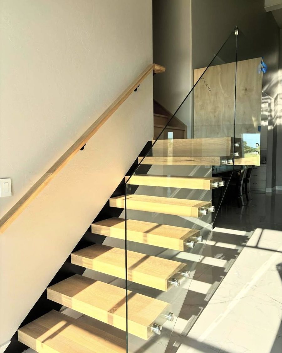 A floating stair with white oak treads cantilevered from the wall, a frameless glass balustrade on standoff fixings, and a wood handrail running the opposite wall