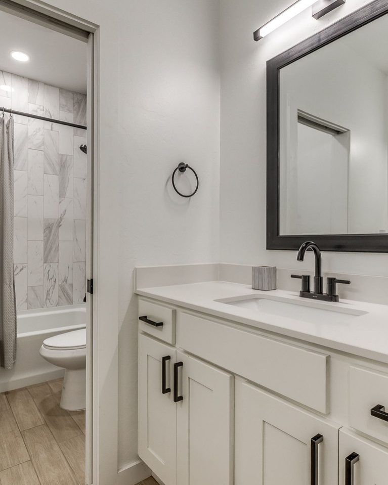 A white shaker vanity with a quartz top, matte black faucet and pulls, and a black framed mirror, with the tub and shower beyond