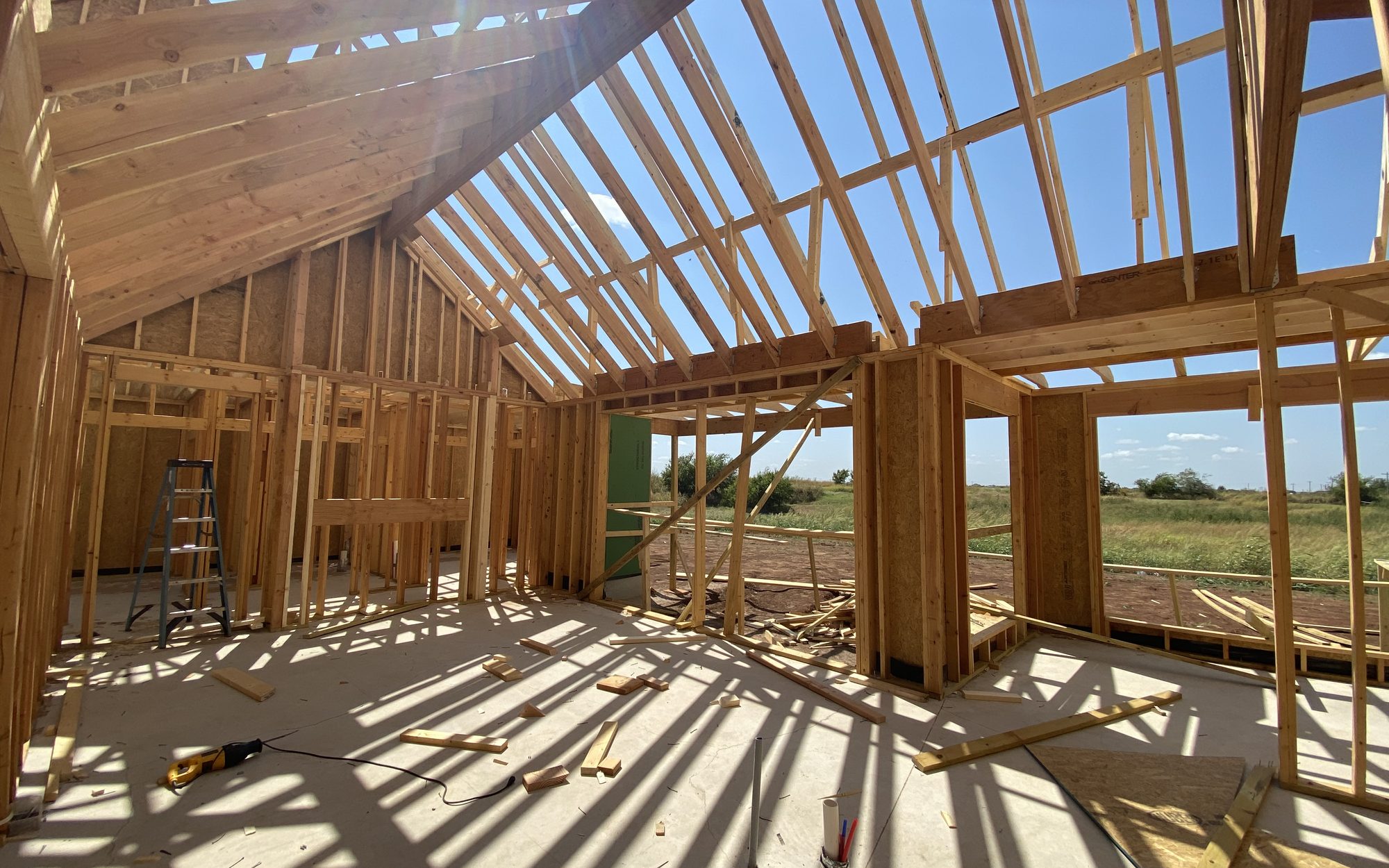 Looking up through the open roof trusses of a house under framing, sunlight casting long striped shadows across the subfloor
