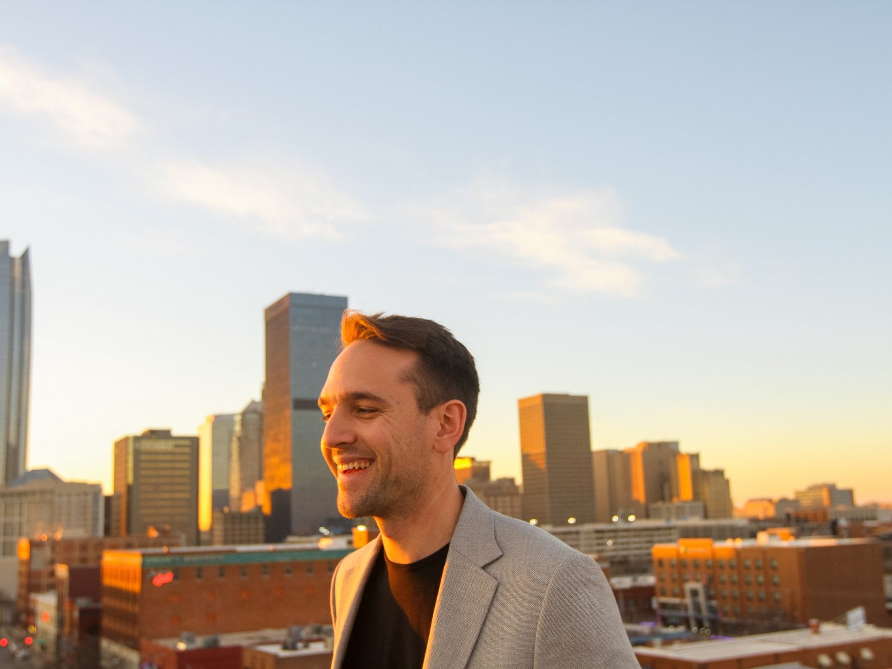 Cayden Ridgeway, owner of Ridgeway Development Group, in a light gray blazer over a black T-shirt, smiling on a rooftop at sunset with the downtown Oklahoma City skyline behind him