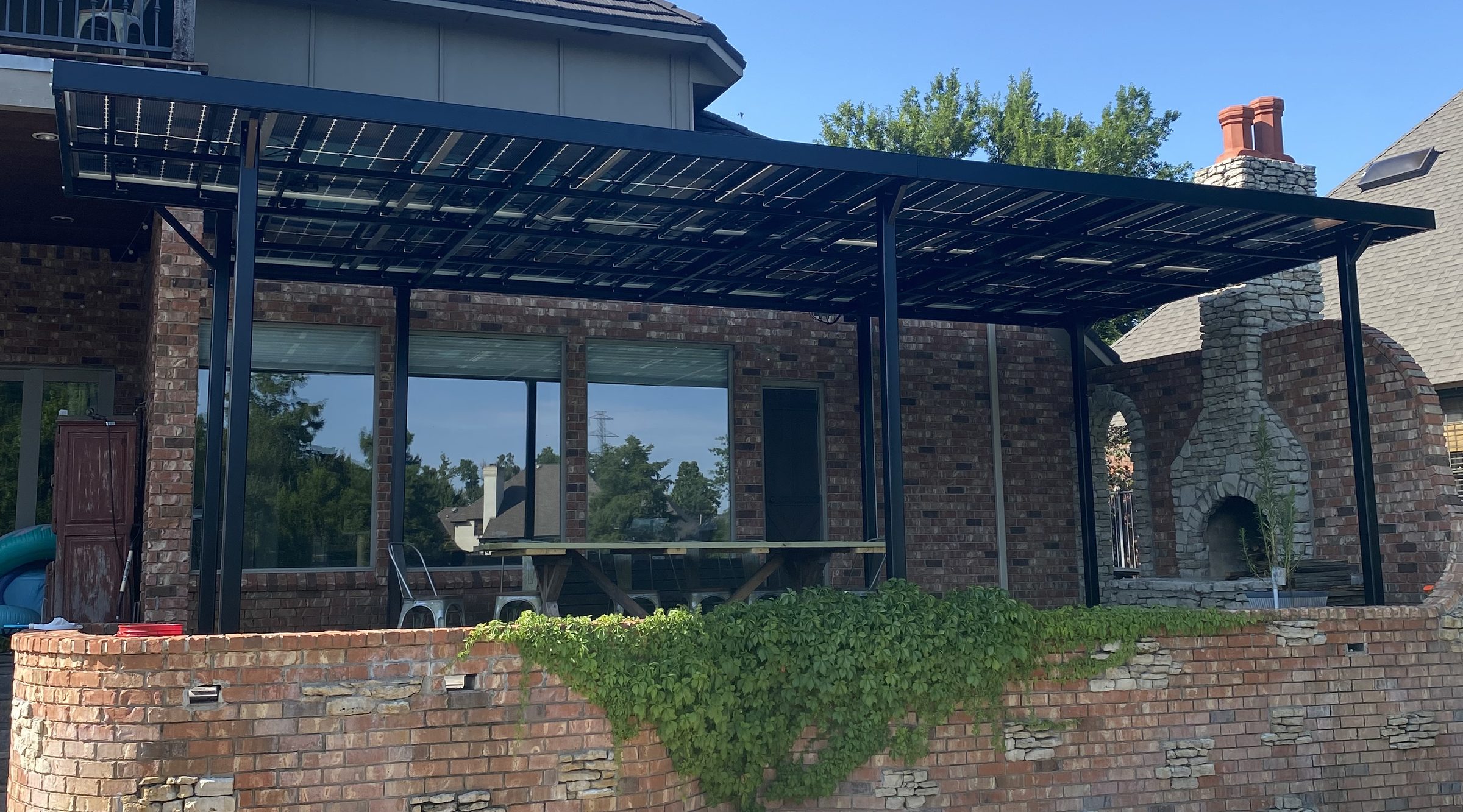 A bi-facial solar pergola on brick columns over a paved terrace, looking out past the outdoor fireplace to the pool and lawn