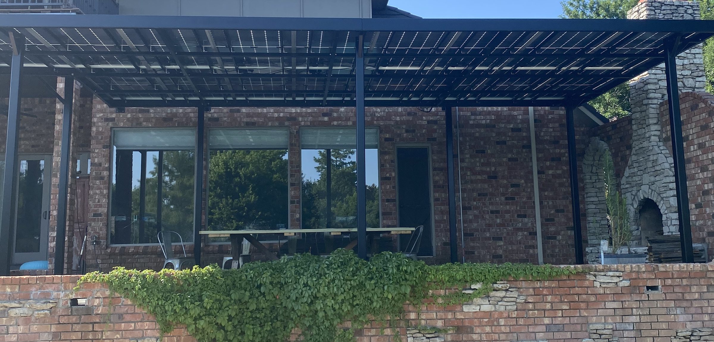 The full run of a bi-facial solar pergola in black steel along the back of a brick house, with the outdoor fireplace at one end