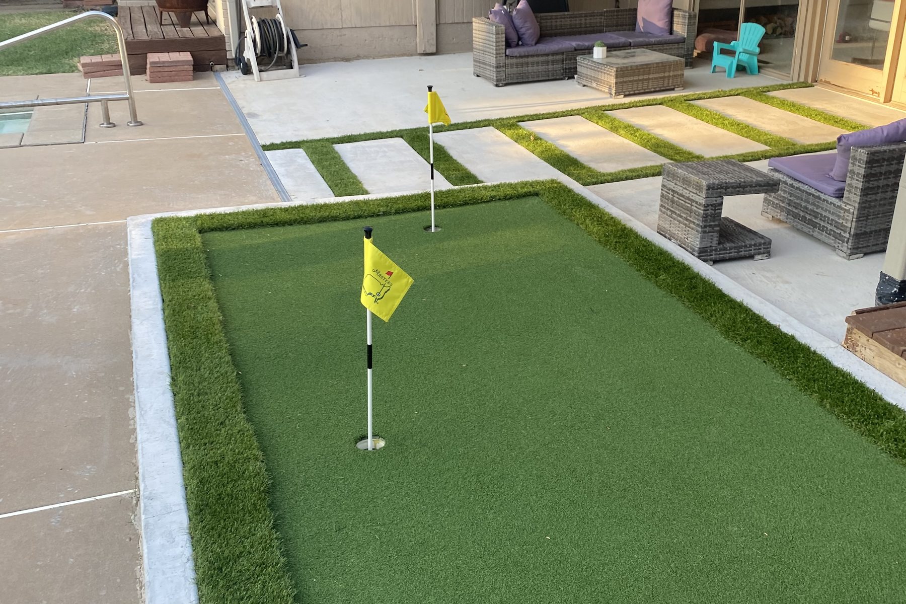 A finished backyard putting green with a flag in the cup, bordered by a poured concrete edge, with the banded terrace alongside it