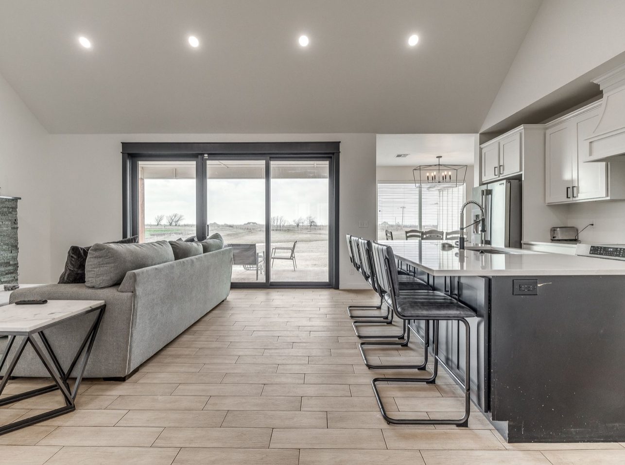 An open living room and kitchen under a vaulted ceiling, a gray sectional facing sliding doors to the patio and a dark island with bar stools