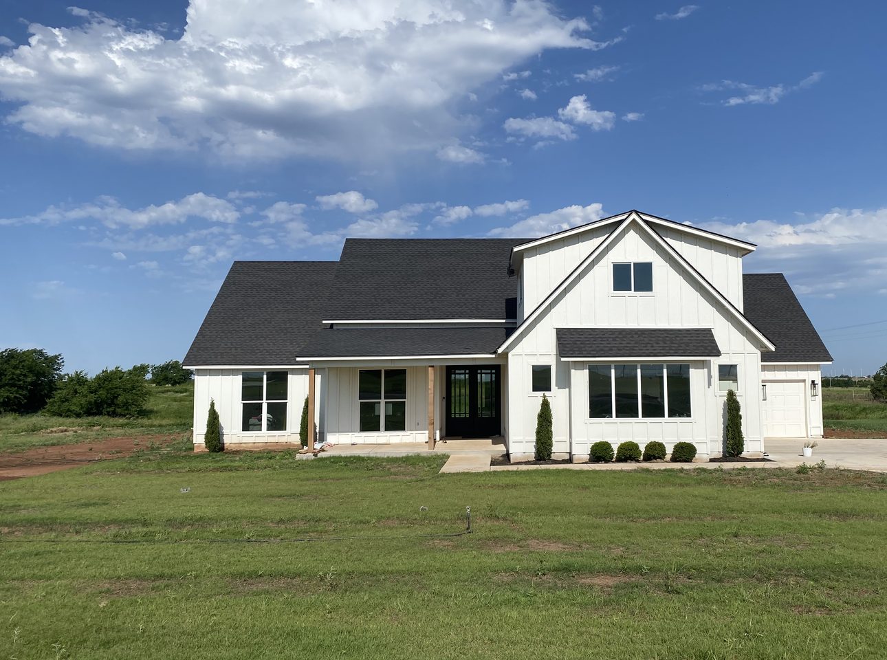 A finished spec home seen from the front on a summer morning, white board and batten under a dark roof, a covered porch on cedar posts and young shrubs along the foundation, on a mown lawn